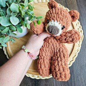 May include: A handmade brown teddy bear with a white muzzle and dark eyes. The bear is made of a textured, plush material. The bear is resting on a round, woven tray. A bracelet with pink and green floral accents is visible.
