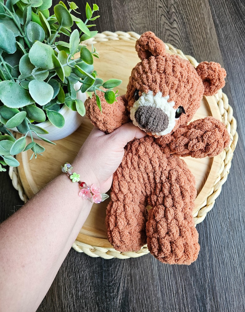 May include: A handmade brown teddy bear with a white muzzle and dark eyes. The bear is made of a textured, plush material. The bear is resting on a round, woven tray. A bracelet with pink and green floral accents is visible.