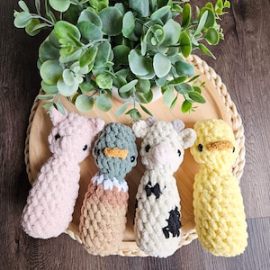 May include: Four crocheted stuffed animals in the shape of a pig, duck, cow, and a bird. The animals are sitting on a wooden tray with a green plant in the background.