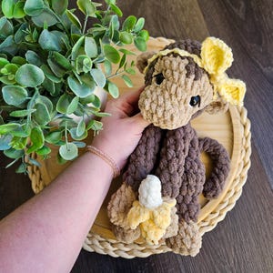 May include: A handmade crochet monkey doll in shades of brown and tan, with a yellow bow and a banana. The plush toy is held in a person's hand, and is sitting on a wooden tray.