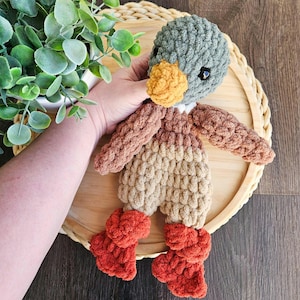 Puede incluir: Un juguete de peluche de pato tejido a crochet con cabeza verde, cuerpo marr&oacute;n y beige y botines naranjas. El pato est&aacute; sentado en una bandeja de madera.