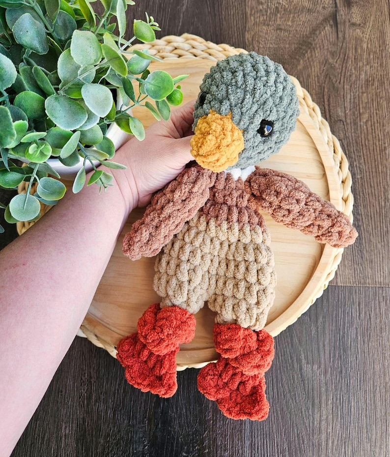 Puede incluir: Un juguete de peluche de pato tejido a crochet con cabeza verde, cuerpo marr&oacute;n y beige y botines naranjas. El pato est&aacute; sentado en una bandeja de madera.
