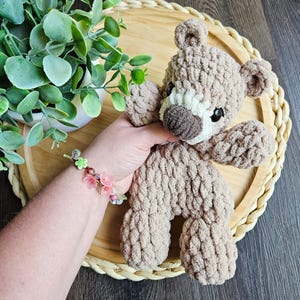 May include: A handmade, light brown crochet teddy bear with a white muzzle and dark brown nose. The bear is sitting on a round, woven tray. A hand holds the bear, and a bracelet with pink flowers is visible.