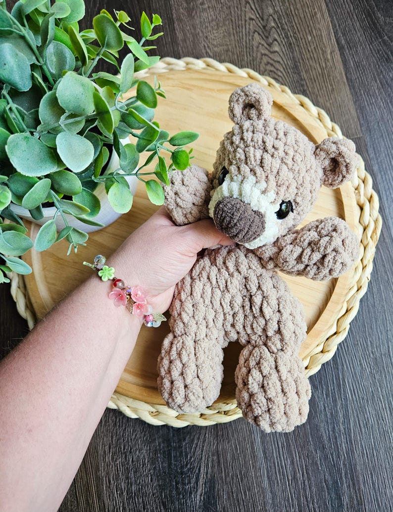 May include: A handmade, light brown crochet teddy bear with a white muzzle and dark brown nose. The bear is sitting on a round, woven tray. A hand holds the bear, and a bracelet with pink flowers is visible.