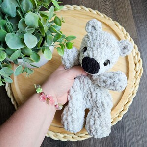 May include: A handmade gray teddy bear with a black nose and small black eyes. The bear is made of a soft, textured yarn. The bear is on a round, woven tray with a green plant in the background.