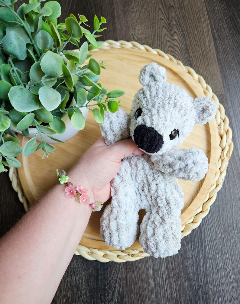 May include: A handmade gray teddy bear with a black nose and small black eyes. The bear is made of a soft, textured yarn. The bear is on a round, woven tray with a green plant in the background.