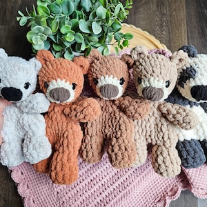 May include: A collection of five handmade crochet teddy bears in various colors, including gray, orange, brown, tan, and a panda-style bear. Each bear has a contrasting snout and black eyes, resting on a pink crocheted blanket.