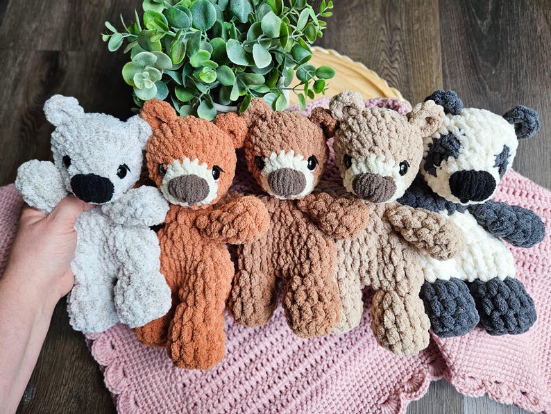 May include: A collection of five handmade crochet teddy bears in various colors, including gray, orange, brown, tan, and a panda-style bear. Each bear has a contrasting snout and black eyes, resting on a pink crocheted blanket.
