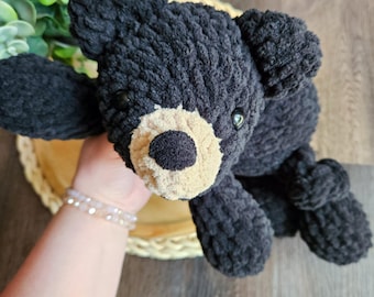 May include: A handmade black bear plush toy with a tan muzzle and small black eyes. The bear is made of a soft, textured yarn and has a cute, cuddly appearance. The bear is lying on a light brown surface.