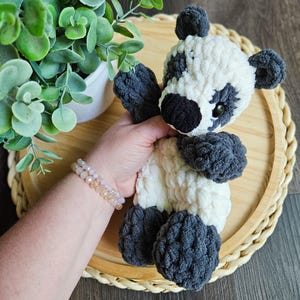May include: A handmade crochet panda bear with a white body and black accents on the ears, eyes, and limbs. The plush toy is soft and cuddly, perfect for children or as a decorative item. The panda is held in front of a wooden tray.