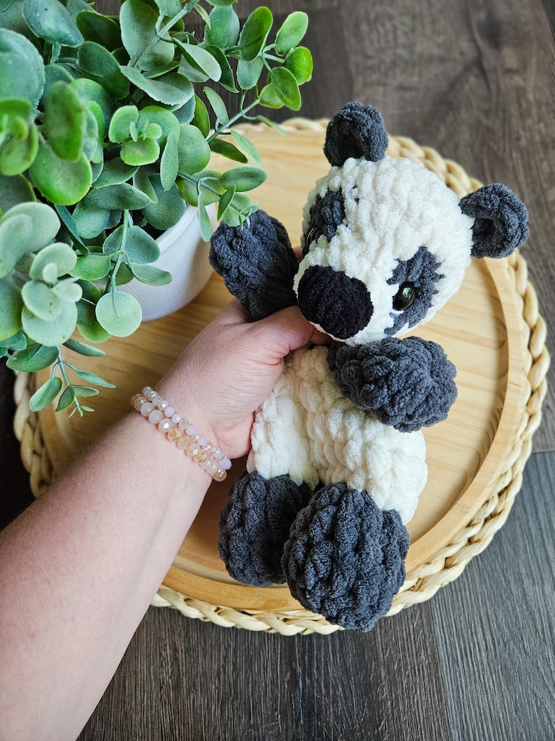 May include: A handmade crochet panda bear with a white body and black accents on the ears, eyes, and limbs. The plush toy is soft and cuddly, perfect for children or as a decorative item. The panda is held in front of a wooden tray.