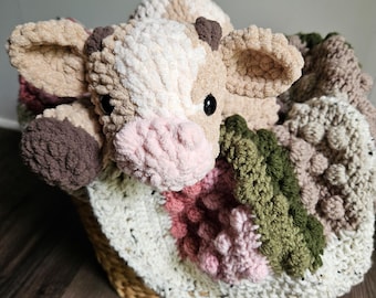 May include: A handmade crochet cow plush toy with a pink nose and black eyes, nestled in a woven basket with a matching blanket. The blanket features shades of white, pink, green, and brown.