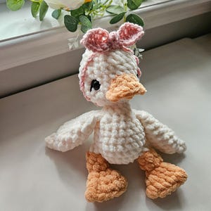 May include: A handmade crochet duck plush toy made from soft white yarn with a textured finish. The duck features a light orange beak and webbed feet, black plastic eyes, and a small pink crocheted bow attached to the top of its head.