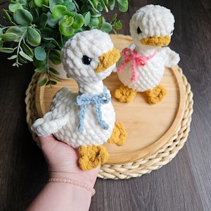 May include: Two handmade white crochet ducklings with yellow beaks and feet. One duckling has a blue bow, the other a pink bow. They are sitting on a wooden tray with a woven edge, near a green plant.