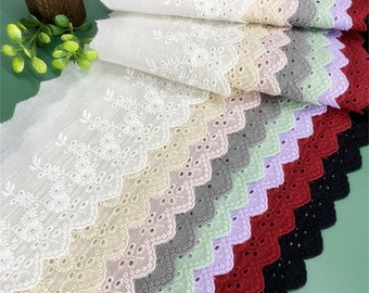 May include: A selection of eyelet lace fabric in a range of colours: white, beige, grey, green, purple, red, and black. The lace has a scalloped edge and floral embroidery. Ideal for sewing and craft projects.