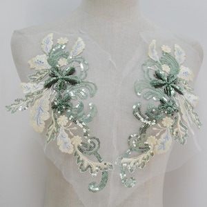 Könnte beinhalten: Zwei grüne und weiße Blumenapplikationen mit Pailletten auf einem weißen Netzstoff. Die Applikationen sind perfekt, um jedem Kleidungsstück oder Bastelprojekt einen Hauch von Eleganz zu verleihen.