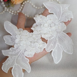 Op de afbeelding: Witte bloemen applicatie met delicate geborduurde details en kleine parel accenten. De applicatie heeft een cluster van bloemen en bladeren, perfect voor het verfraaien van kleding of accessoires. Het ontwerp wordt in een hand gehouden.