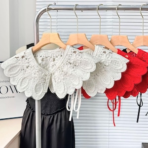 Op de afbeelding: Een collectie kanten kragen in wit, rood en zwart, hangend aan houten kleerhangers. De kragen hebben ingewikkelde bloempatronen en striksluitingen. De kragen worden getoond op een metalen rek tegen een neutrale achtergrond.