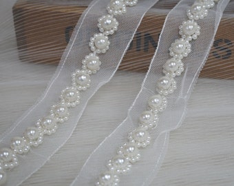 May include: Ivory-colored ribbon with a delicate floral design. Each flower is composed of small, round, faux pearls, creating a refined and elegant trim. The ribbon's sheer fabric adds a touch of lightness, perfect for embellishments.