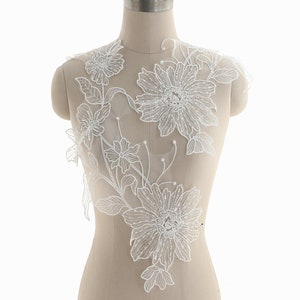 Könnte beinhalten: Weiße florale Spitzenapplikation mit Perlendetails. Die Spitze zeigt ein zartes Blumenmuster mit Blättern und Blüten. Die Applikation ist perfekt, um jedem Kleidungsstück einen Hauch von Eleganz zu verleihen.