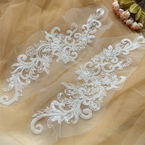 Könnte beinhalten: Zwei weiße Perlen-Spitzenapplikationen mit floralem Muster. Die Applikationen befinden sich auf einem beigefarbenen Tüllstoff.
