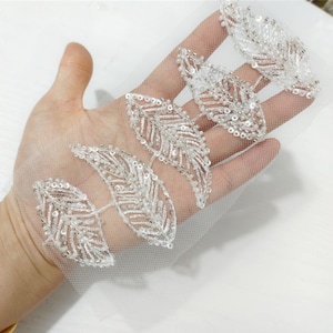 May include: White beaded leaf trim on a sheer white mesh fabric. The trim features a repeating pattern of three leaves.