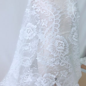May include: White lace fabric with a floral pattern. The lace has a delicate, intricate design with a slightly frayed edge.