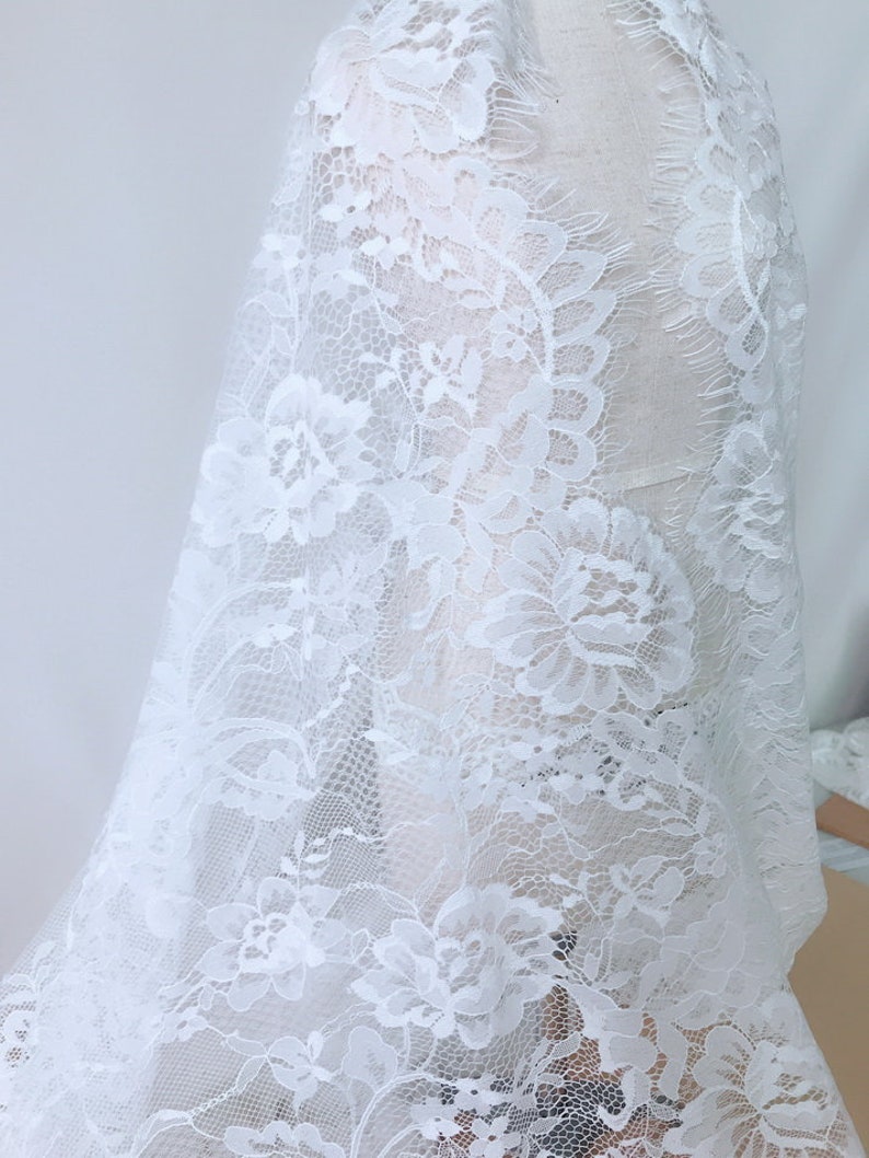 May include: White lace fabric with a floral pattern. The lace has a delicate, intricate design with a slightly frayed edge.