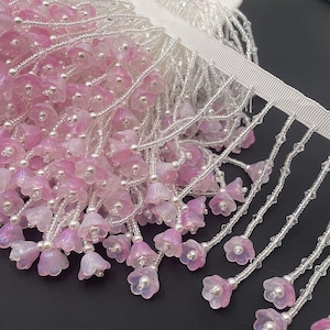 May include: Close-up of a decorative trim with pink and white flower-shaped beads. The beads are strung on clear strands, with a white ribbon base. The trim has a delicate, elegant appearance.