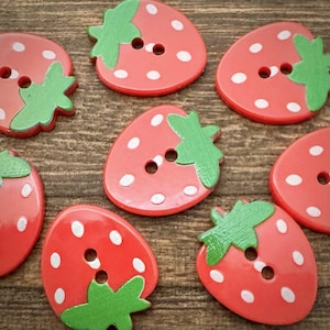 May include: A collection of strawberry-shaped buttons, each featuring a bright red base with white polka dots and a green leaf detail at the top. Each button has two holes in the center for attachment, showcasing a smooth, glossy finish throughout.