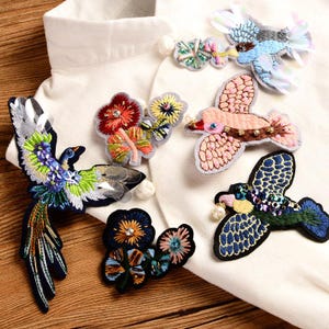 May include: A collection of embroidered patches featuring birds and floral motifs. The patches include a long-tailed bird with green and blue thread, a pink bird, a blue bird with sequins, and two floral clusters with beads and colorful stitching.