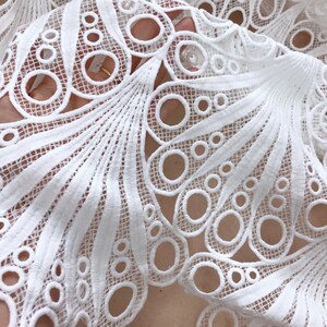 May include: Close-up of white lace fabric with a detailed, openwork design. The lace features a pattern of circles and fan-like shapes, creating a delicate and intricate texture. The fabric is likely used for clothing or decorative purposes.