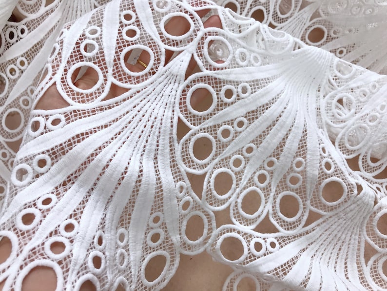 May include: Close-up of white lace fabric with a detailed, openwork design. The lace features a pattern of circles and fan-like shapes, creating a delicate and intricate texture. The fabric is likely used for clothing or decorative purposes.