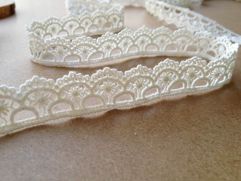 May include: Ivory-colored lace trim with a scalloped edge. The trim is made of a delicate, woven material and is arranged in a wavy pattern. This decorative trim is suitable for various craft projects, including sewing and scrapbooking.