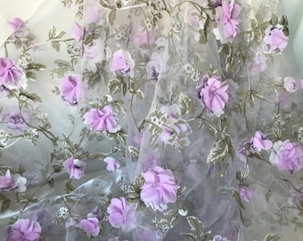 May include: Sheer fabric with a floral design. The fabric features lavender-colored 3D flowers and green and white leaves. The flowers are scattered across the fabric, creating a delicate and romantic look. The fabric is ideal for creating dresses, skirts, or other decorative items.