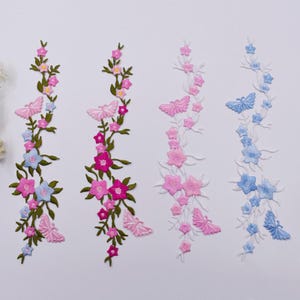 May include: Four vertical embroidered appliques featuring floral vines and butterflies. Two appliques have green stems with pink and blue flowers and butterflies. Two appliques have white stems with pink and blue flowers and butterflies.
