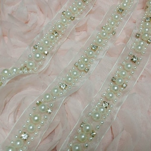 May include: Two strands of white pearl and rhinestone trim on a pink background. The trim is made of a sheer fabric with a scalloped edge.