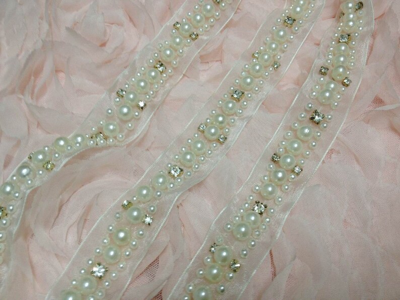 May include: Two strands of white pearl and rhinestone trim on a pink background. The trim is made of a sheer fabric with a scalloped edge.