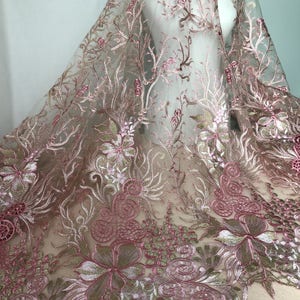 May include: Delicate pink floral embroidered fabric with intricate detailing. The sheer material features a repeating pattern of flowers, leaves, and swirling designs in shades of pink, gold, and white. The fabric is draped over a mannequin.