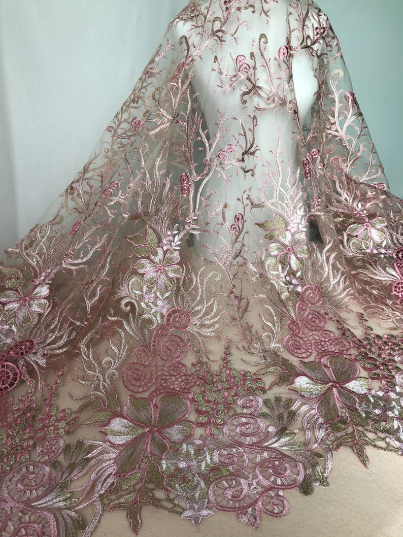 May include: Delicate pink floral embroidered fabric with intricate detailing. The sheer material features a repeating pattern of flowers, leaves, and swirling designs in shades of pink, gold, and white. The fabric is draped over a mannequin.