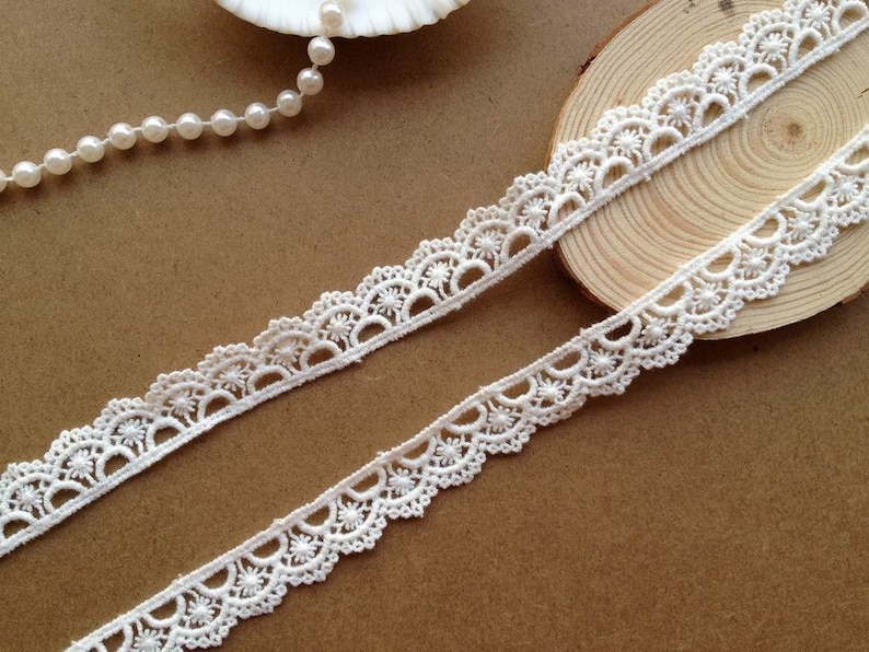 May include: Two strips of off-white lace with a scalloped edge and floral details. The lace is displayed on a brown surface, with a seashell and a string of pearls in the background. The lace is likely used for sewing, crafting, or embellishing.