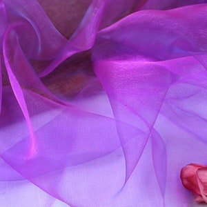 May include: Purple sheer fabric with a soft, flowing texture. The fabric is draped over a white surface with a single pink rose in the bottom right corner.