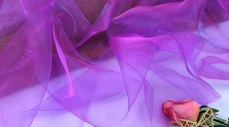 May include: Purple sheer fabric with a soft, flowing texture. The fabric is draped over a white surface with a single pink rose in the bottom right corner.