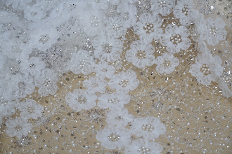 以下が含まれることがあります： 大きな3Dの花のアップリケと小さな虹色のビーズが付いた白いレース生地。生地は繊細で透き通った質感です。