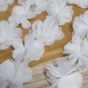 Puede incluir: Primer plano de adornos florales de tela blanca. Las flores están en capas con delicados pétalos y un respaldo de malla. Las flores están dispuestas sobre una superficie de madera marrón claro, creando una estética suave y aireada. Ideal para manualidades y embellecimiento.