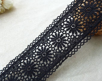 May include: Close-up of black lace trim featuring a floral daisy design. The lace has a scalloped edge and is made of a delicate, open-weave fabric. The trim is ideal for sewing, crafting, and adding a decorative touch to garments or home decor projects.