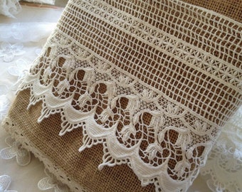 May include: A textured brown burlap fabric runner featuring a wide band of intricate cream-colored lace trim. The lace displays a detailed floral pattern with scalloped edges and a geometric grid design, layered over the coarse woven natural fiber material.