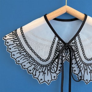 May include: A white sheer collar with black embroidered lace trim. The collar has a tie closure in the front.