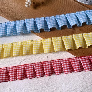 May include: Three rows of gingham fabric trim in blue, yellow, and red. The trim is pleated, creating a ruffled effect. The fabric has a classic checkered pattern, perfect for crafts and sewing projects.