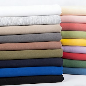 Pode incluir: Pilhas de tecido em várias cores, incluindo branco, cinza, bege, cinza-marrom, verde-oliva, azul marinho, azul e preto. Outra pilha apresenta tons pastel de rosa, vermelho, verde, amarelo e roxo. O tecido parece ser macio e texturizado.